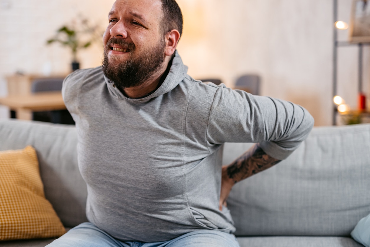 Person with pained expression sitting on home sofa with both hands on lower back